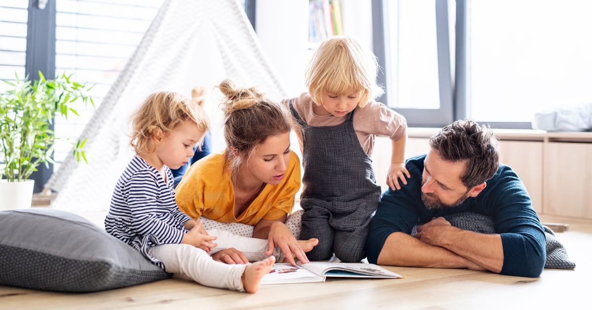 Young mom and dad laying on ground reading story book to two toddlers