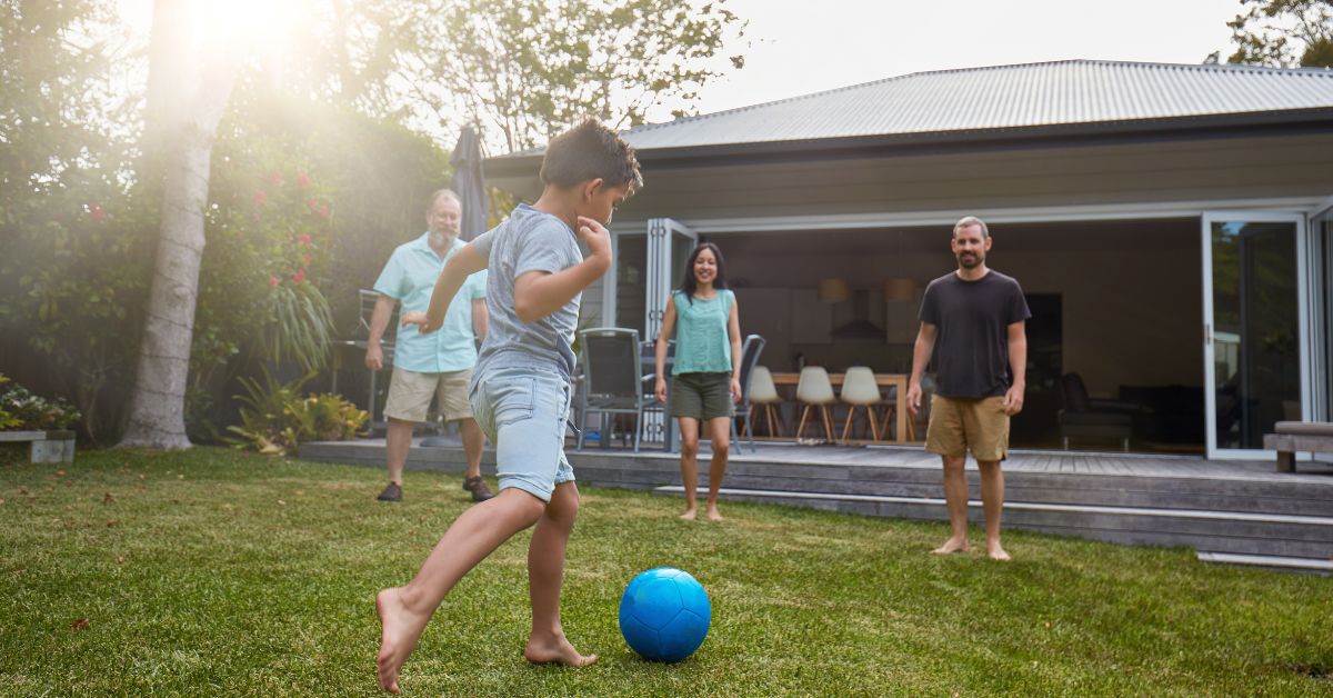 Young boy kicking blue soccer ball in backyard with 3 adults smiling and encouraging
